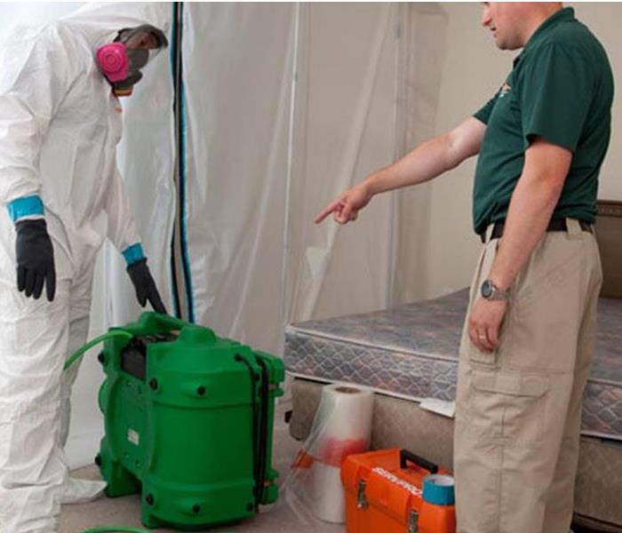 SERVPRO tech dressed in full protection suit, a manager pointing to big green bucket and a bed in the background