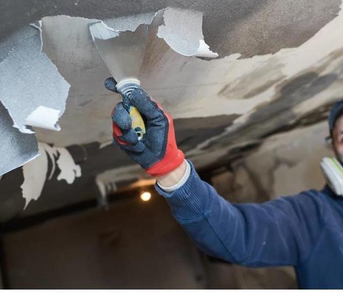 A worker in a mask scrapes soot-covered paint off of a ceiling